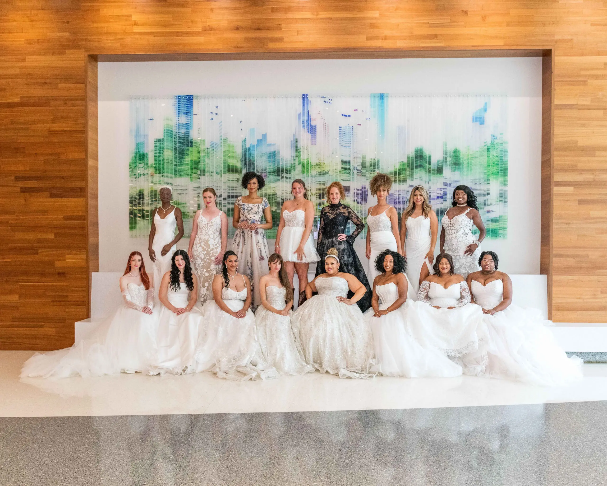 A diverse group of sixteen models wearing a variety of wedding gowns—ranging from classic ballgowns to modern fitted silhouettes—pose in front of a vibrant cityscape mural at the Houston Bridal Extravaganza, photographed by Nathan Hamilton of In a Lifetime Photography.