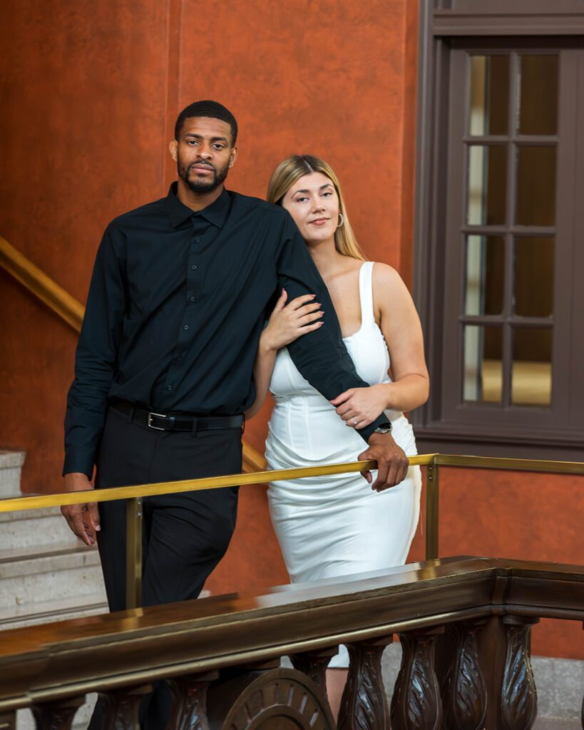 A sophisticated couple leaning against a dark wood railing on the mezzanine balcony of the Julia Ideson Library during their Houston engagement session.